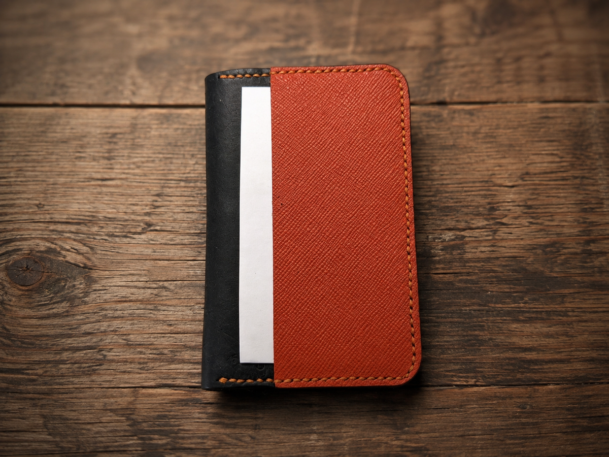 A minimalist card holder wallet with a textured orange-red front cover and a black side panel, shown on a rustic wooden background.