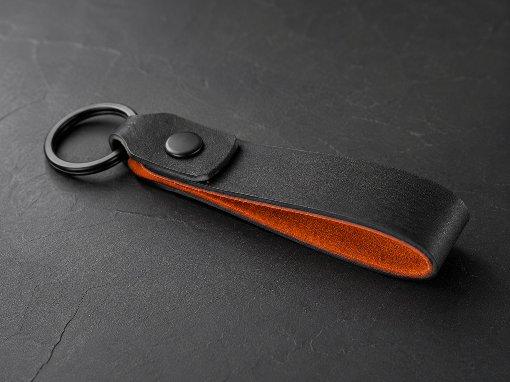 A black leather keychain pouch with a black metal key ring and snap button, featuring a vibrant orange interior lining, set against a dark textured slate background.