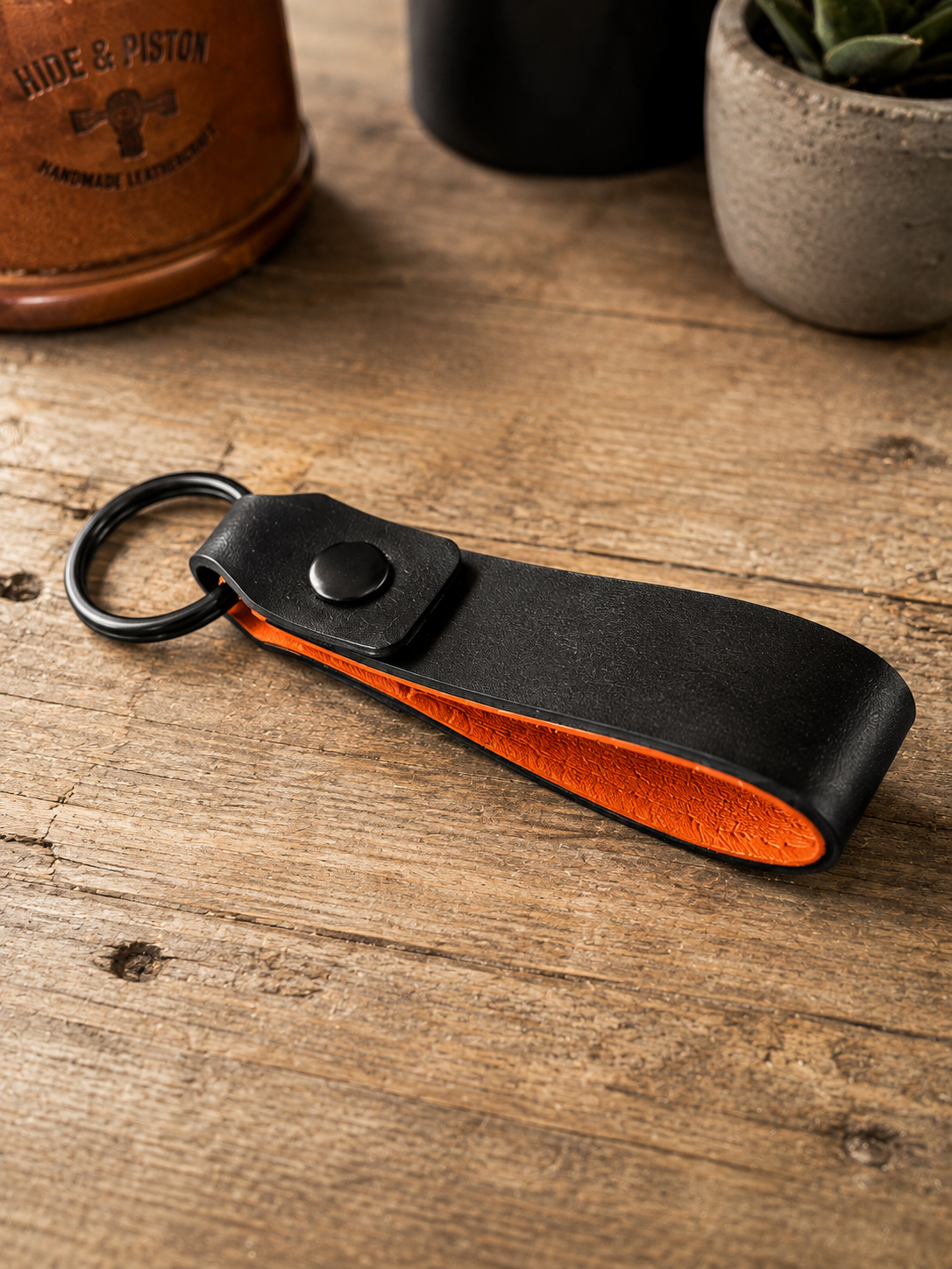 A black leather keychain with a bright orange interior lining and a black metal key ring, resting on a rustic wooden tabletop.