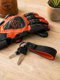 An orange and black leather motorcycle glove with an embroidered logo, displayed on a light wood tabletop next to a black leather key fob with keys.