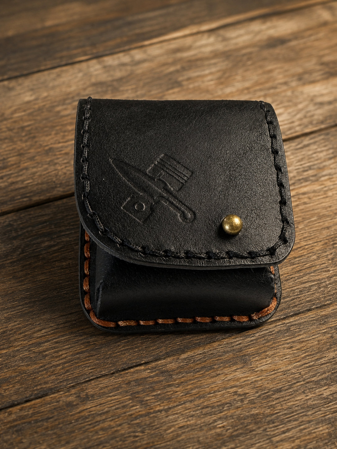 A small black leather card holder or wallet with a snap button closure, placed on a rustic wooden surface. The leather has visible stitching, an embossed logo on the front, and a small brass snap button.