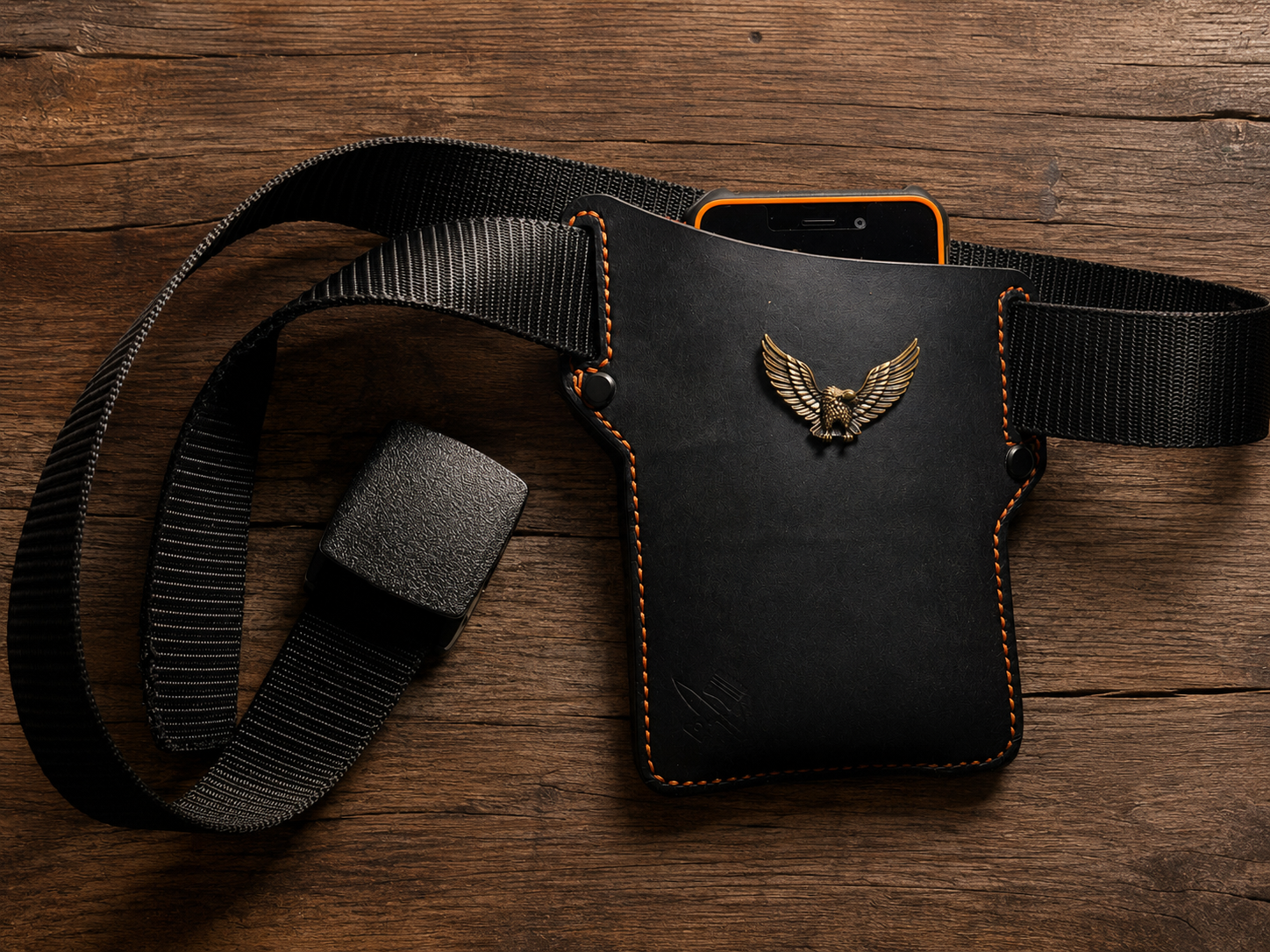 A black leather phone pouch with a gold eagle emblem, attached to a black webbing belt, displayed on a rustic wooden surface with a smartphone tucked inside.