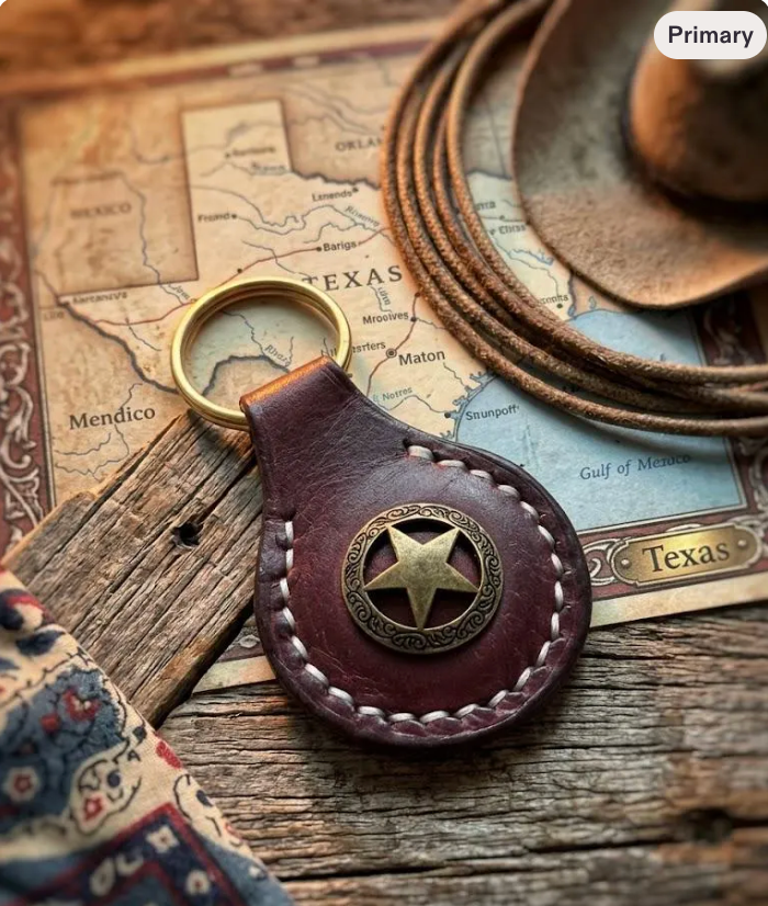 A western-style leather keychain with a round metal key ring and a star emblem, styled on a rustic wooden surface with a map of Texas, coiled ropes, and a hat.