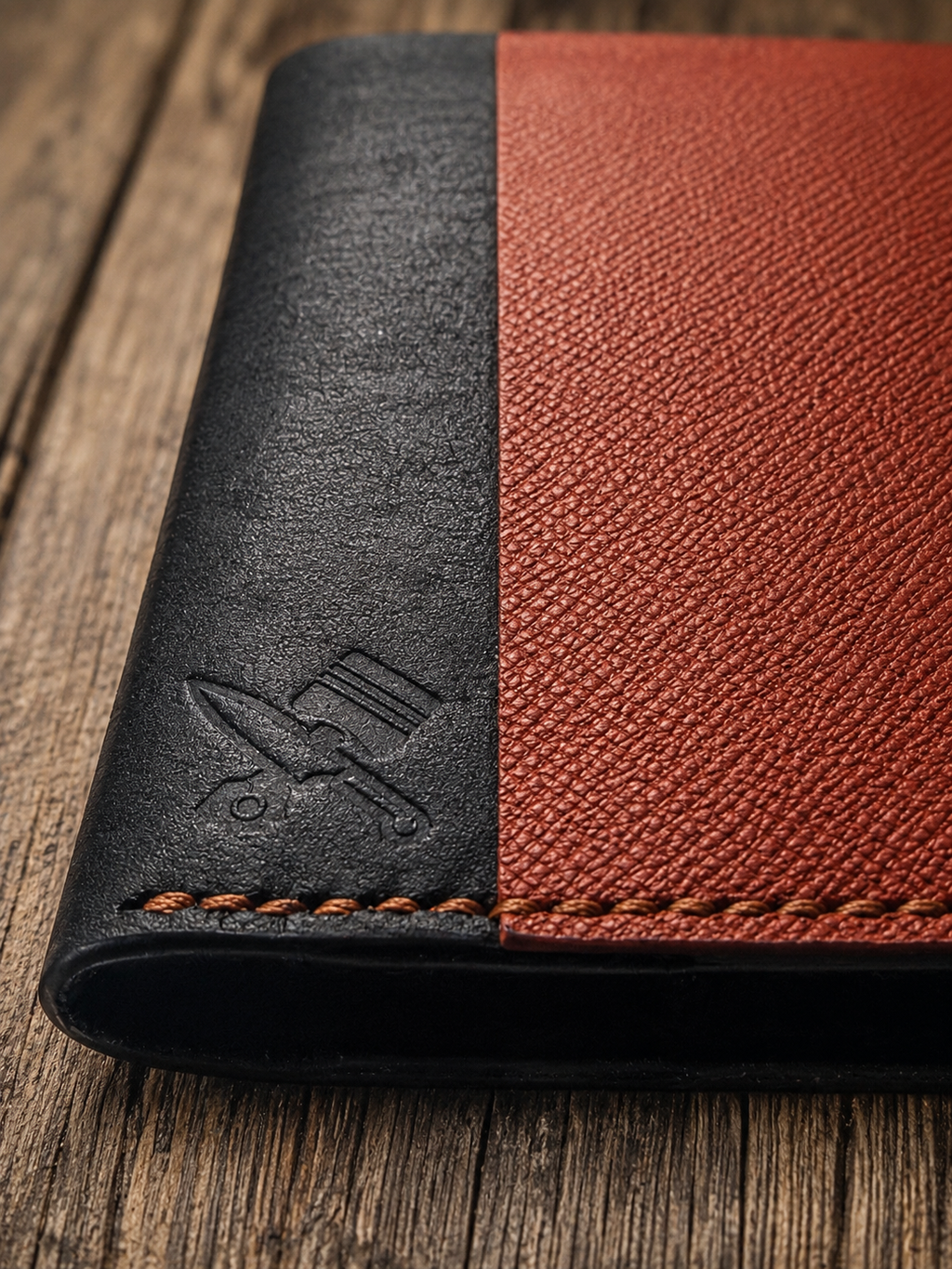 A close-up of a leather wallet or folio with a split design: one half is matte black with an embossed logo, and the other half is textured reddish-brown. The item has visible orange stitching and is photographed on a rustic wooden surface.