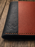 A close-up of a leather wallet or folio with a split design: one half is matte black with an embossed logo, and the other half is textured reddish-brown. The item has visible orange stitching and is photographed on a rustic wooden surface.