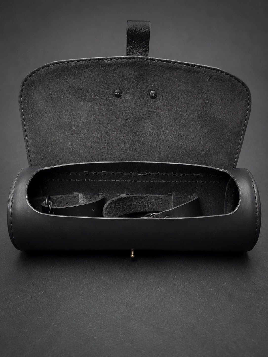 A black leather cylindrical case with an open hinged lid, shown on a dark gray background. Inside the case, a black strap or belt is partially visible.