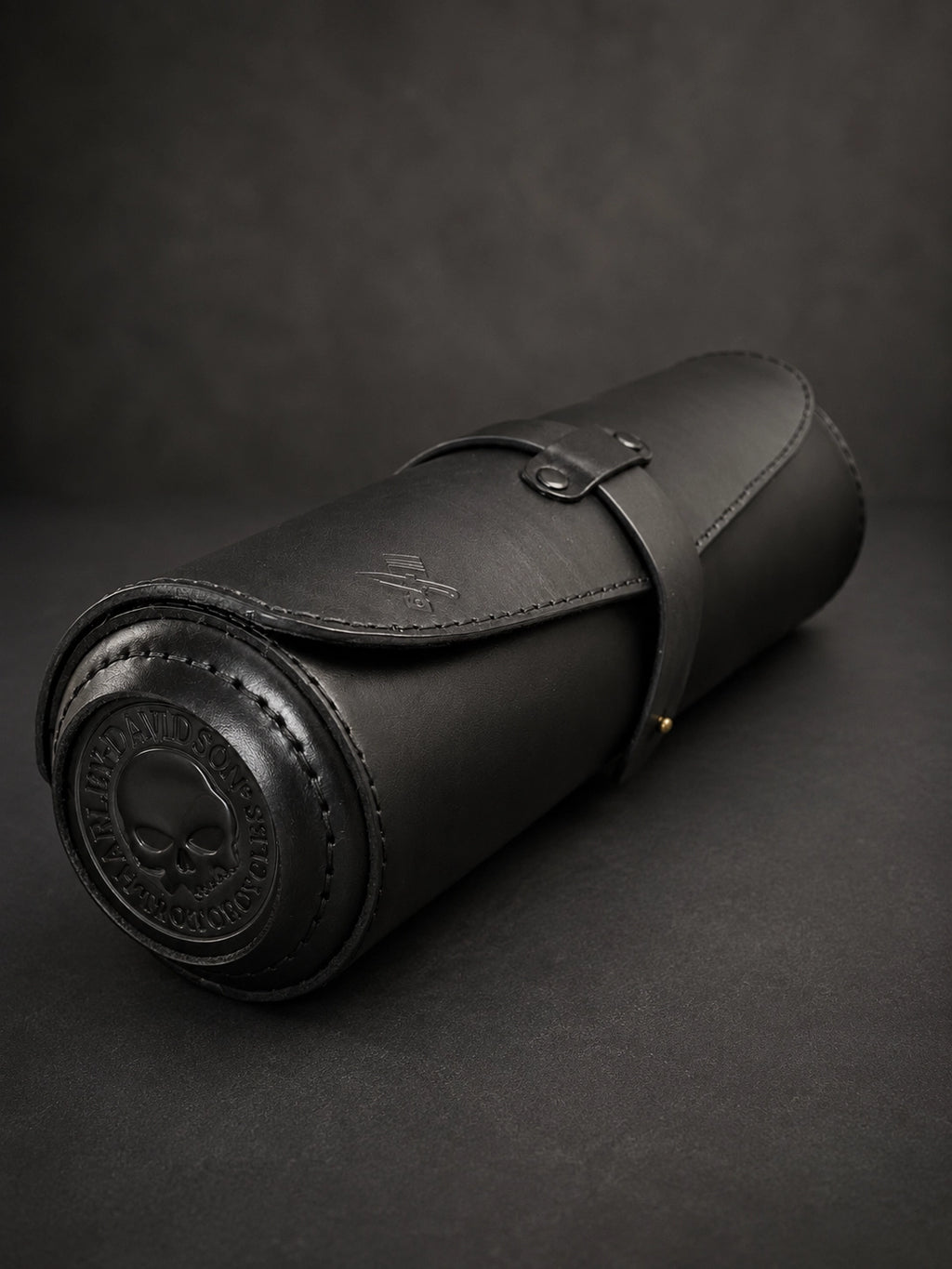 A black leather cylindrical roll-up case with a strap closure and embossed skull detailing on the end, photographed on a dark textured background.