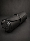 A black leather cylindrical roll-up case with a strap closure and embossed skull detailing on the end, photographed on a dark textured background.