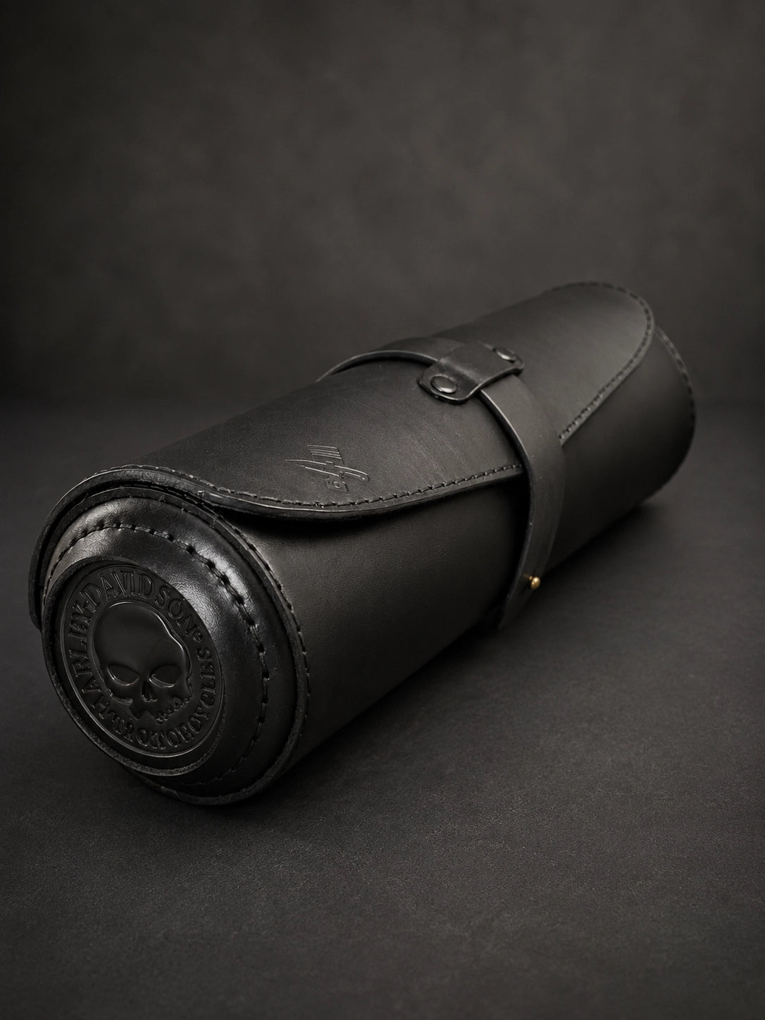 A black leather cylindrical roll-up case with a strap closure and embossed skull detailing on the end, photographed on a dark textured background.