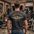 A man viewed from behind wearing a dark heather gray t-shirt with a gold graphic print on the back that reads 'HIDE & PISTON' and 'Built to Ride' with a motorcycle-themed emblem. He is standing in a brick garage with motorcycles in the background.