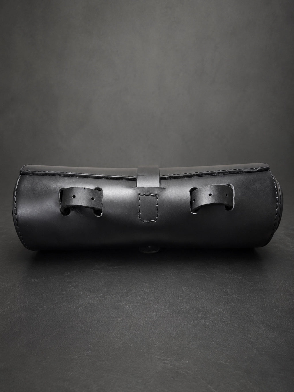 A black leather cylindrical case with two front strap closures and a stitched front flap, photographed on a dark gray studio background.