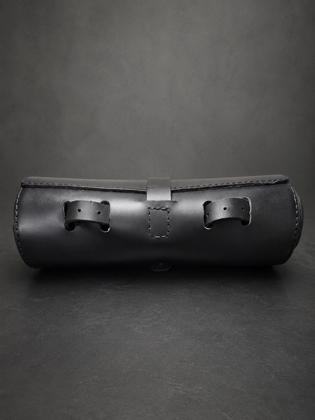 A black leather cylindrical case with two front strap closures and a stitched front flap, photographed on a dark gray studio background.